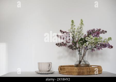 Moment de tranquillité avec tasse de café, plateau en osier. Bouquet floral de branches de lilas violettes et blanches en fleurs dans un vase en verre. Petit déjeuner printanier Banque D'Images