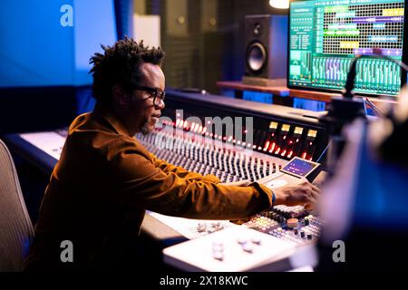 Producteur de musique afro-américain composant des pistes dans la salle de contrôle, éditant des chansons et ajustant les paramètres de volume avec les boutons du tableau de bord. Ingénieur du son appuyant sur les faders et les boutons. Banque D'Images