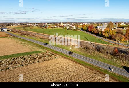 La fin de l'après-midi, le soleil baigne une scène rurale paisible, avec un buggy tiré par des chevaux et des véhicules sur une route de campagne, soulignant le mélange de tradition et de modernité. Banque D'Images