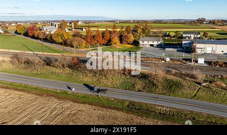 Un buggy amish serein parcourt une route à Lancaster, PA, flanqué par les couleurs vives de l'automne et de vastes terres agricoles, un témoignage du riche patrimoine agricole de la région. Banque D'Images