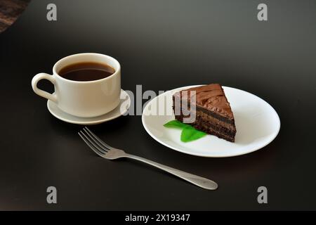 Un morceau de gâteau de couche de chocolat avec des feuilles de menthe, une fourchette et une tasse de café noir sur une soucoupe sur un fond noir. Gros plan. Banque D'Images