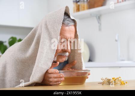 Homme mûr avec serviette faisant inhalation de vapeur à table dans la cuisine Banque D'Images