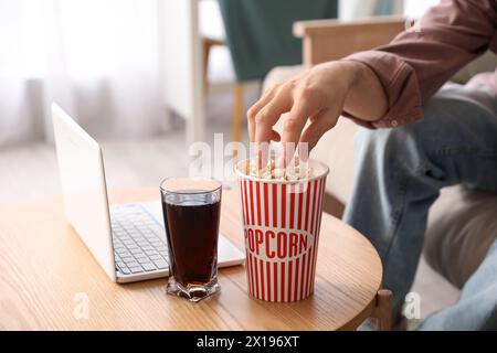Main masculine prenant pop-corn de la table avec ordinateur portable et verre de cola dans la chambre Banque D'Images