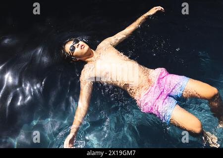 Un adolescent asiatique portant des lunettes de soleil flottant dans l'eau claire de la piscine à l'extérieur à la maison, l'air détendu Banque D'Images