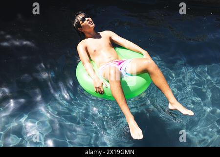 Un adolescent asiatique portant des lunettes de soleil se relaxant sur un flotteur vert dans une piscine à l'extérieur à la maison Banque D'Images