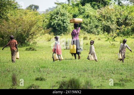 Dans une forêt africaine, une mère ougandaise pieds nus porte deux cruches à eau tandis que son plus jeune enfant regarde la caméra. Les enfants, pieds nus également, accompagnement Banque D'Images