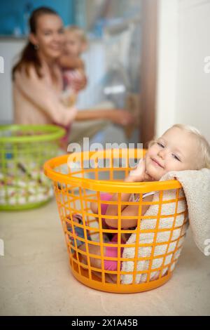 Bébé heureux, portrait et se détendre avec maman dans le panier à linge pour l'enfance amusante, le jeu ou la journée de corvée à la maison. Jeune petite fille avec sourire dans le seau pendant Banque D'Images