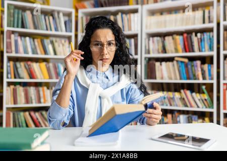 Une bibliothécaire sérieuse d'âge moyen avec des lunettes examine attentivement un livre tout en organisant les étagères de la bibliothèque, entourée d'une vaste collection de livres. Banque D'Images