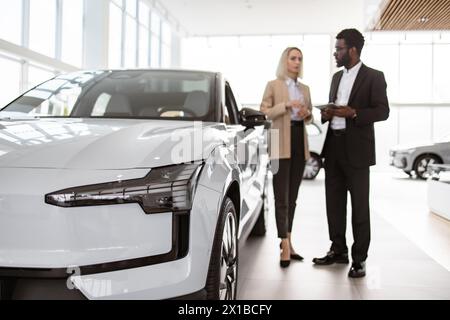 Le consultant masculin africain fournit des informations sur la nouvelle voiture en utilisant la tablette à la cliente féminine caucasienne à la concession automobile. Femme d'affaires réussie en voiture Banque D'Images