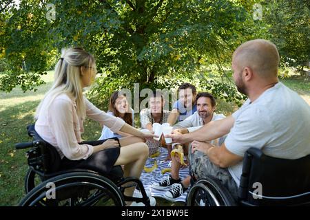 Un groupe diversifié d'amis, y compris des personnes utilisant des fauteuils roulants, partageant un pique-nique joyeux à l'extérieur par une journée ensoleillée, soulignant l'inclusivité et les amis Banque D'Images
