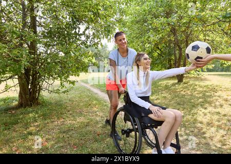 Une scène joyeuse dans un parc représentant deux amis, dont l'un est une personne en fauteuil roulant, souriant tout en interagissant avec un ballon de soccer par une journée ensoleillée. Banque D'Images