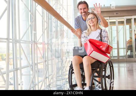 Un moment joyeux comme un homme salue chaleureusement son amie, une femme en fauteuil roulant, avec un coffret cadeau rouge, symbolisant l'amitié et le soutien dans un accessibl Banque D'Images