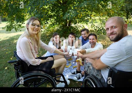 Groupe d'amis heureux interagissant avec une personne qui utilise un fauteuil roulant lors d'un pique-nique en plein air dans un parc. Banque D'Images