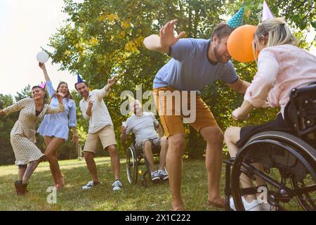 Un groupe diversifié d'amis, y compris une personne qui utilise un fauteuil roulant, célèbrent à l'extérieur avec joie et chapeaux de fête. Banque D'Images