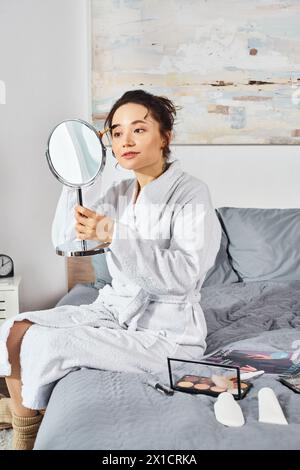 Une femme brune dans un peignoir blanc est assise sur un lit, examinant attentivement quelque chose avec un miroir entouré de cosmétiques. Banque D'Images