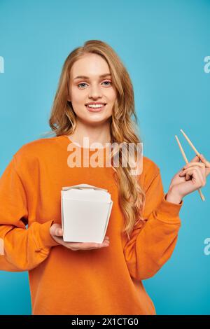 Une belle femme aux cheveux blonds tient délicatement la boîte de nourriture et les baguettes, savourant la cuisine asiatique. Banque D'Images
