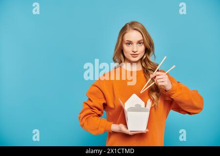 Une belle femme blonde tient délicatement des baguettes et une boîte de délicieux plats asiatiques. Banque D'Images