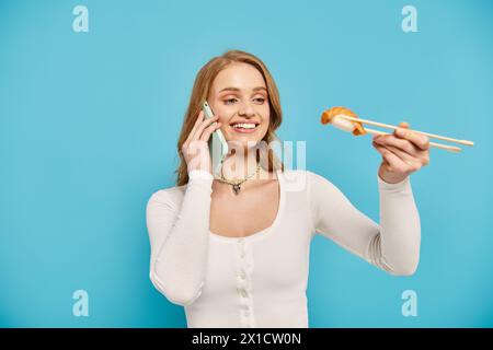 Une femme blonde tient délicatement des baguettes tout en discutant au téléphone, appréciant la cuisine asiatique. Banque D'Images