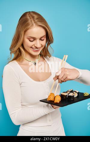 Une belle femme aux cheveux blonds tient délicatement une assiette de sushi et de baguettes. Banque D'Images