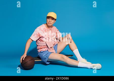 Un jeune homme élégant et beau dans une tenue tendance assis sur le sol, tenant un ballon de basket, sur fond bleu. Banque D'Images