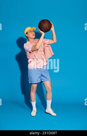 Jeune homme élégant tient en toute confiance un ballon de basket dans sa main droite, respirant athlétisme et fraîcheur sur fond bleu. Banque D'Images