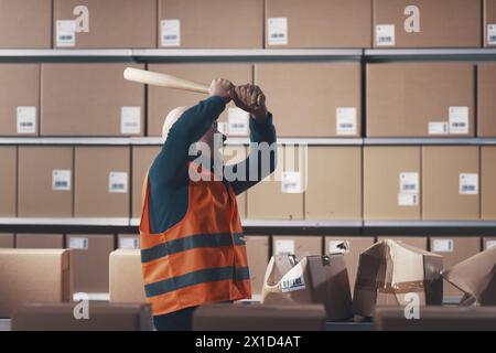 Ouvrier d'entrepôt rebelle fou brisant des boîtes en carton avec une batte de baseball Banque D'Images