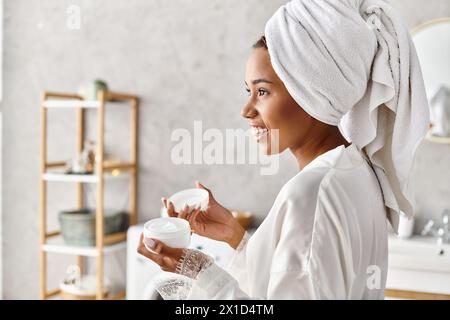 Une femme, avec une serviette sur la tête, tenant un pot avec de la crème dans sa salle de bain, profitant d'un moment de détente et de tranquillité. Banque D'Images