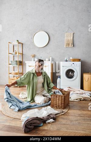 Une femme avec des tresses afro est assise sur le sol devant une machine à laver, s'attaquant à son linge dans la salle de bain. Banque D'Images