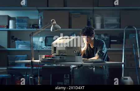 Mec nerd intelligent assis à la réception et utilisant un vieil ordinateur dans le garage la nuit Banque D'Images