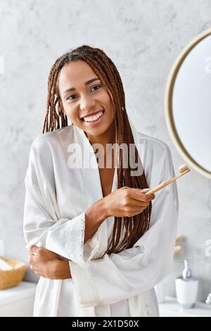 Femme afro-américaine en peignoir avec tresses afro se brossant les dents dans un miroir de salle de bain moderne. Banque D'Images