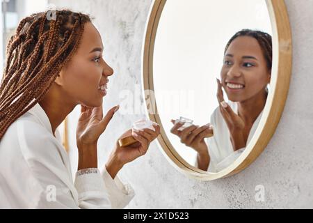 Une femme afro-américaine avec des tresses afro appliquant de la crème dans une salle de bain moderne tout en portant un peignoir. Banque D'Images