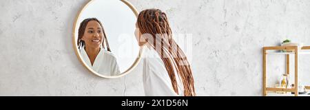 Une femme afro-américaine avec des tresses afro regarde son reflet dans un miroir dans une salle de bain moderne, contemplant beauté et hygiène. Banque D'Images