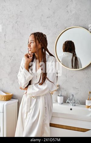 Femme afro-américaine avec des tresses afro se tient dans un peignoir devant le miroir de salle de bain moderne. Banque D'Images