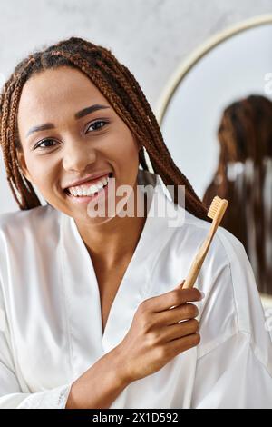 Une femme avec des dreadlocks dans un peignoir tient une brosse à dents dans une salle de bain moderne, en se concentrant sur la beauté et l'hygiène. Banque D'Images