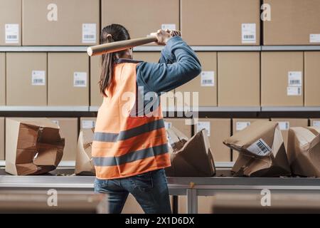 Ouvrier d'entrepôt rebelle en colère brisant des boîtes sur la bande transporteuse avec une batte sur le lieu de travail, vue arrière Banque D'Images