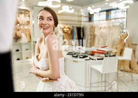 Une jeune mariée brune et belle se tient en toute confiance devant un miroir dans un magasin de vêtements, essayant différentes robes blanches pour son jour spécial. Banque D'Images