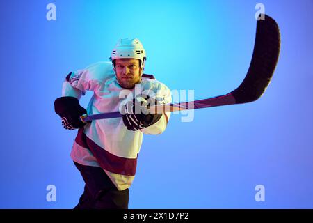 Homme concentré, joueur de hockey avec bâton montrant sa motivation pour gagner, posant sur fond bleu dégradé dans la lumière néon Banque D'Images