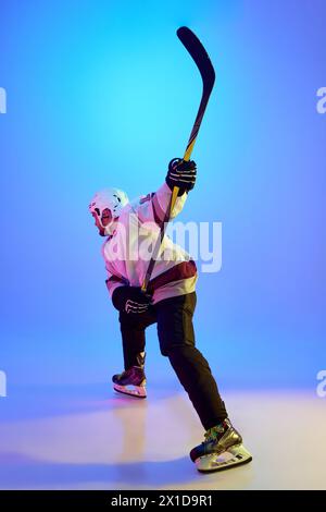 Homme ambitieux, joueur de hockey posant avec un bâton, montrant sa détermination sur fond bleu dégradé dans la lumière néon Banque D'Images