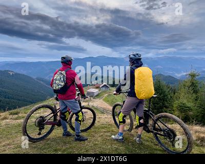 Deux hommes cyclistes conduisant des vélos électriques à l'extérieur. Vue arrière des touristes masculins se reposant sur le sommet de la colline, appréciant le beau paysage de montagne, portant casque et sac à dos. Concept de loisirs actifs. Banque D'Images