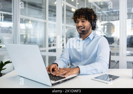 Représentant masculin joyeux du service clientèle avec micro-casque/oreillette travaillant sur ordinateur portable dans un environnement de bureau lumineux et contemporain. Communication et assistance professionnelles. Banque D'Images