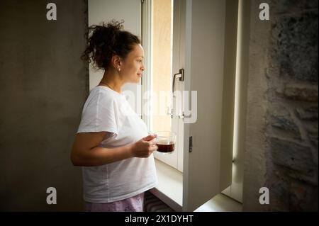 Belle jeune femme adulte en pyjama debout près de la fenêtre avec une tasse de boisson chaude, en admirant la belle vue depuis la fenêtre. Banque D'Images