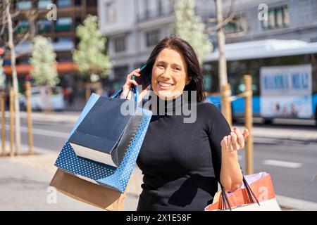 élégante femme d'âge moyen parlant sur téléphone intelligent commandant un taxi avec des sacs à provisions Banque D'Images