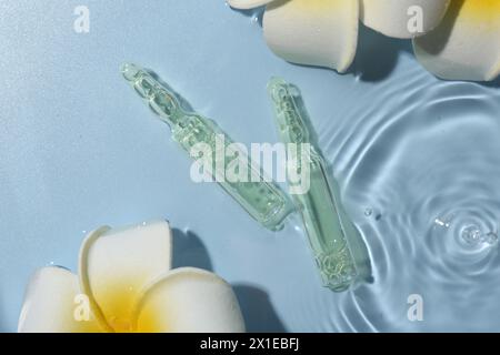 Ampoules de soins de la peau et belles fleurs de plumeria dans l'eau sur fond bleu clair, pose à plat Banque D'Images