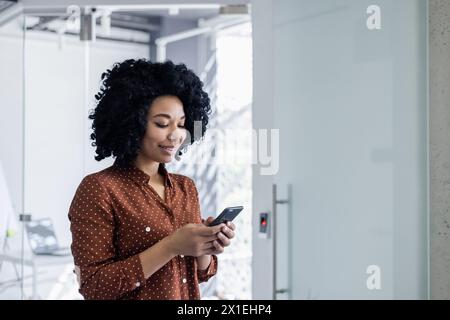 Une femme afro-américaine professionnelle dans une élégante blouse à pois utilise son smartphone dans un environnement de bureau lumineux et moderne, à la recherche de contenu et concentré. Banque D'Images