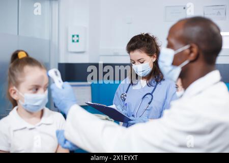 Femme en bleu gommage écrivant des notes tandis que l'homme noir dans une blouse de laboratoire vérifie la température de l'enfant caucasien. L'image montre un médecin masculin examinant le patient avec l'aide d'une infirmière. Banque D'Images