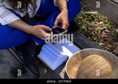 Jeune femme latino-américaine (33 ans) méconnaissable, profite de l’arrivée du printemps pour s’asseoir sur un banc de parc et écrire des messages texte sur son ph cellulaire Banque D'Images