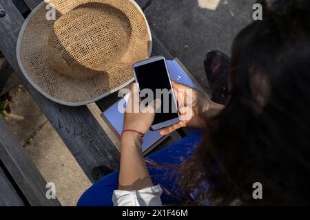 Jeune femme latino-américaine (33 ans) méconnaissable, profite de l’arrivée du printemps pour s’asseoir sur un banc de parc et écrire des messages texte sur son ph cellulaire Banque D'Images