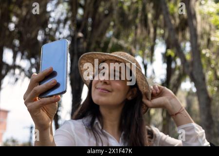 Au printemps, une jeune femme latino-américaine (33 ans) est assise sur un banc de parc souriante tout en utilisant son téléphone portable pour un autoportrait. en utilisant son phon cellulaire Banque D'Images