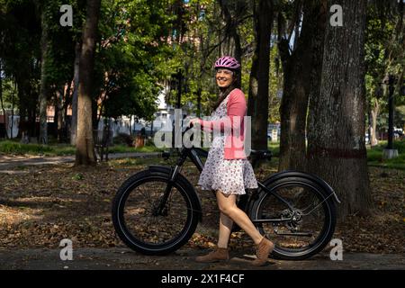 Une jeune femme latino-américaine (33 ans) monte son vélo électrique dans le parc au printemps, portant un casque et le soleil illumine son visage. Concept signifie Banque D'Images