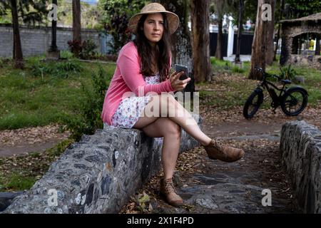 Une jeune femme latino-américaine (33 ans) est assise dans le parc en envoyant des SMS. Elle porte un chapeau, une robe fleurie et un pull rose. Backgrou électrique pour bicyclettes Banque D'Images